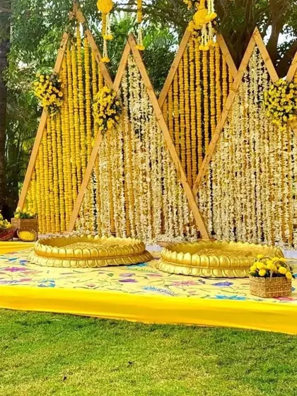 A beautiful outdoor Haldi setup featuring a backdrop of hanging marigold and jasmine garlands against triangular wooden frames. This is perfect for a rustic or garden-themed event.
