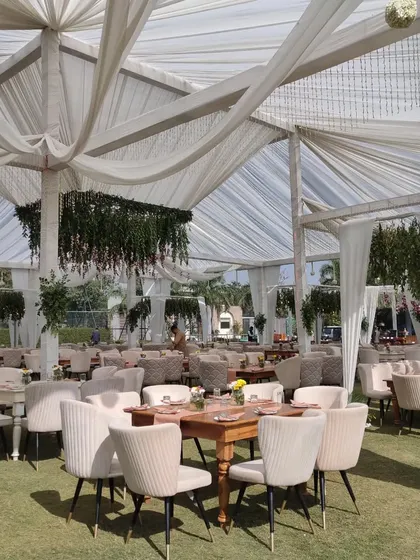 A chic lounge seating arrangement for a daytime outdoor event. The combination of modern upholstered chairs, wooden tables, and draped white canopies creates a comfortable and stylish space for guests to relax.