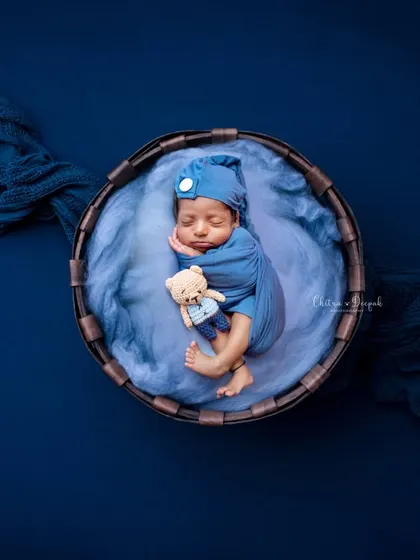 A classic newborn pose, with the baby swaddled snugly in blue and tucked into a rustic basket with a tiny teddy bear. The deep blue background creates a rich, portrait-like effect.