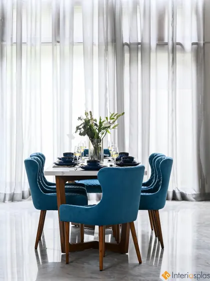 A beautiful dining area with blue upholstered chairs and a simple, elegant table setting. The large windows with sheer curtains create a bright and airy atmosphere.