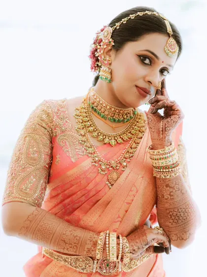 A detailed portrait of a bride in a peach-colored saree, highlighting her intricate temple jewellery and henna-adorned hands.