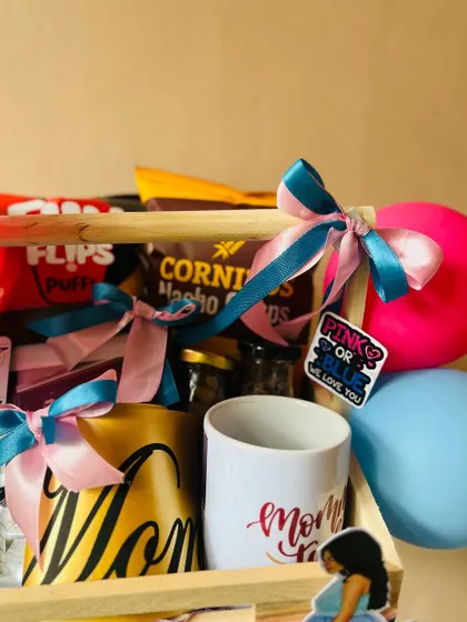 A close-up of a mom-to-be gift hamper. This view highlights the personalized mugs and snack selection, tied with festive pink and blue ribbons.