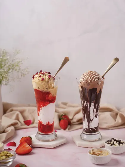A pair of tall, decadent milkshakes, one strawberry and one chocolate. The styling with fresh fruit and chocolate chips highlights the ingredients and makes the drinks look irresistible.