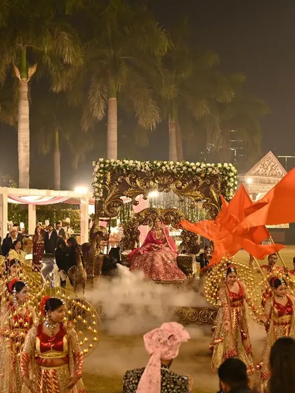 A truly grand bride entry. The bride is carried on an ornate palanquin, surrounded by a large troupe of dancers with flags and props, creating a scene of royal splendor.