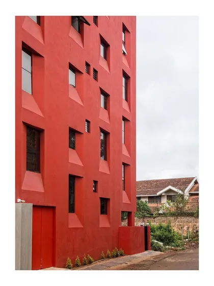 The building's red facade contrasts with the traditional tiled roof of a neighboring house, highlighting the project's contemporary approach in a dense, evolving suburb.