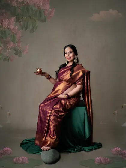 A regal portrait of a mom-to-be in a deep maroon silk saree. The painterly background with lotus flowers adds an artistic and serene touch.