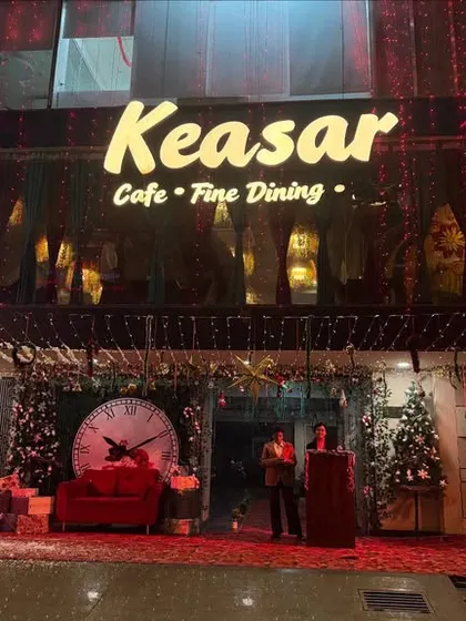 A large-scale Christmas decoration for the entrance of Keasar Cafe. The setup includes a photo booth with a giant clock, a Christmas tree, and gift boxes.