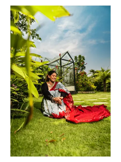 A candid, happy moment in the garden. The vibrant red and grey saree stands out against the green grass and blue sky, creating a picture of pure joy.