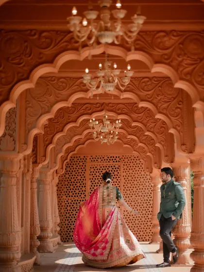 The bride twirls in her beautiful lehenga while her groom watches, captured in a grand hallway with intricate arches and chandeliers.