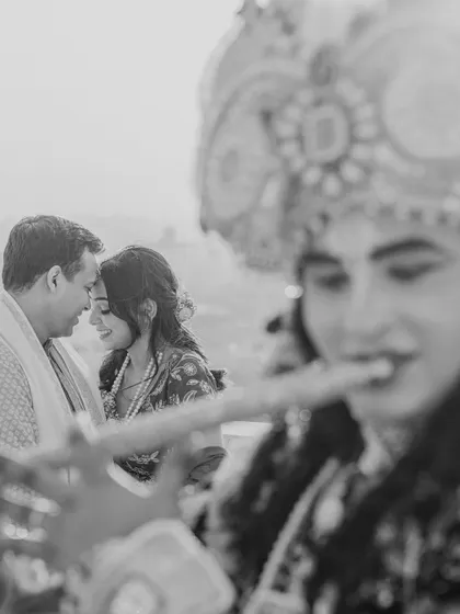 An artistic black and white shot capturing an intimate moment between the couple, with a performer dressed as Krishna playing the flute in the foreground. This creative composition is both unique and deeply romantic.