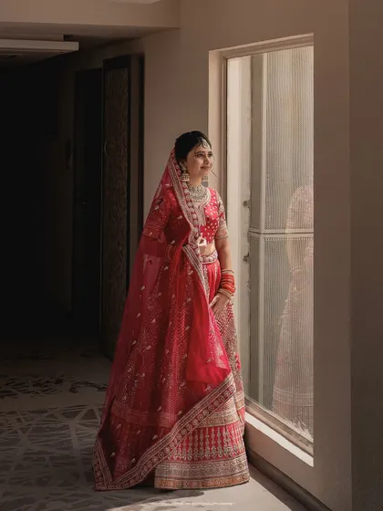 A moment of quiet anticipation. The bride looks out the window, her reflection captured beside her, creating a thoughtful and artistic portrait in her beautiful red lehenga.