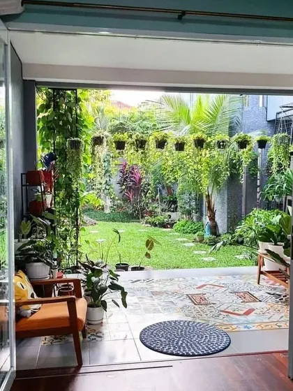 A view from inside looking out to a cozy patio and garden. The design uses patterned tiles on the patio floor, comfortable seating, and a variety of hanging and potted plants to create a seamless indoor-outdoor living experience.
