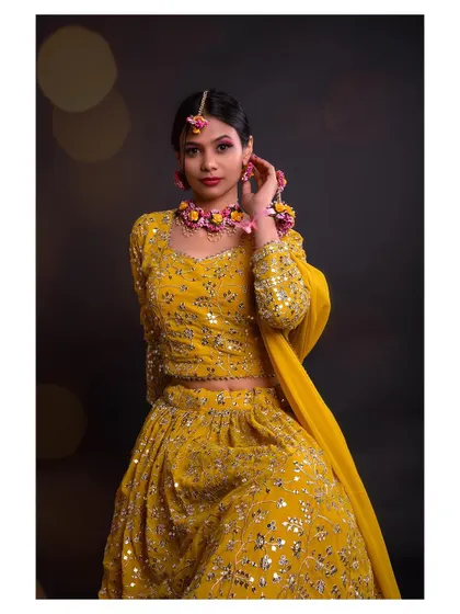 A studio portrait of a woman in a vibrant yellow lehenga and floral jewelry, perfect for a Haldi or Mehendi look. The dark background makes the colors pop.
