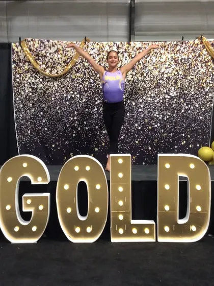 An Xcel Gold athlete poses in front of the "GOLD" sign at regionals in Colorado.