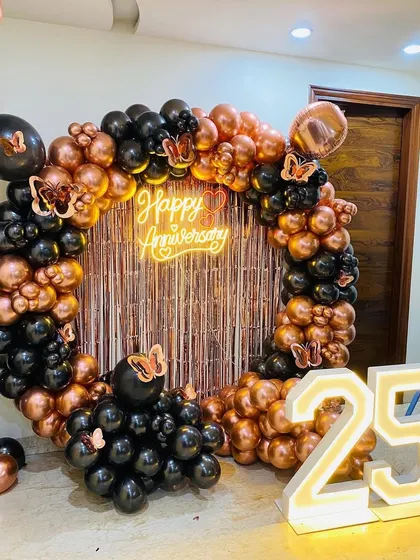 Celebrating a 25th anniversary with a stylish black and rose gold theme. This decor features a full ring of balloons, a shimmering fringe backdrop, a "Happy Anniversary" neon sign, and large light-up numbers to mark the milestone.