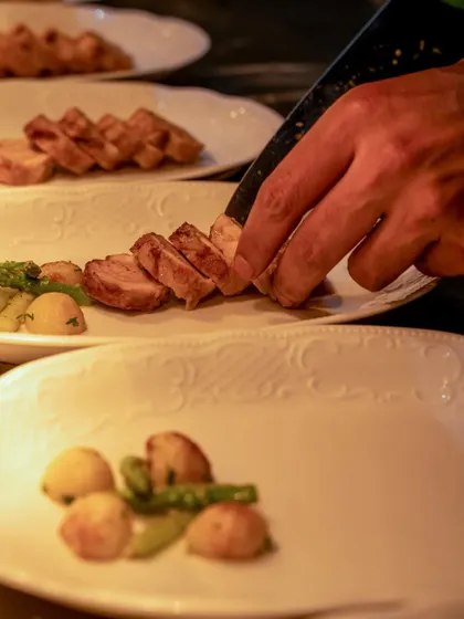 A chef's hand slicing meat and arranging it on a plate with potatoes and asparagus.