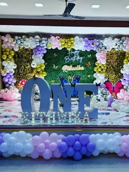 A stunning butterfly-themed first birthday stage for Rithvika. The design combines a green hedge wall with gold shimmer curtains, a large "ONE" prop, and a beautiful balloon arrangement in purple, white, and gold.