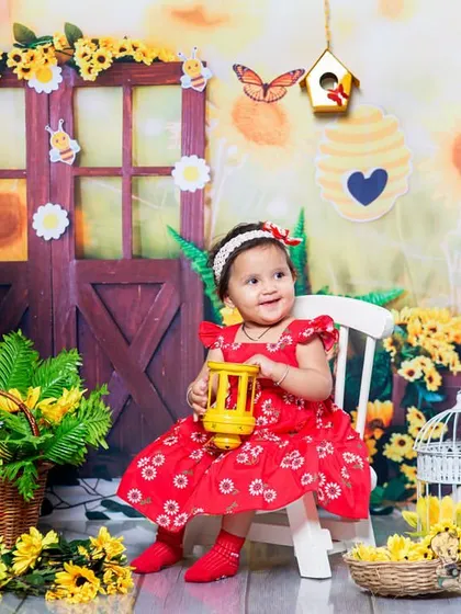 A toddler in a red dress sits in a vibrant sunflower and bee-themed garden set. The bright yellows and her happy smile create a sunny and cheerful portrait.