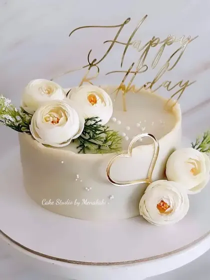 A pretty no-fondant birthday cake in all white. This elegant design is adorned with white flowers and a golden heart accent for a clean, classic, and romantic feel.