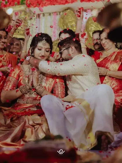 The groom blesses his bride with flower petals, a beautiful and intimate moment from a traditional South Indian wedding, surrounded by family.
