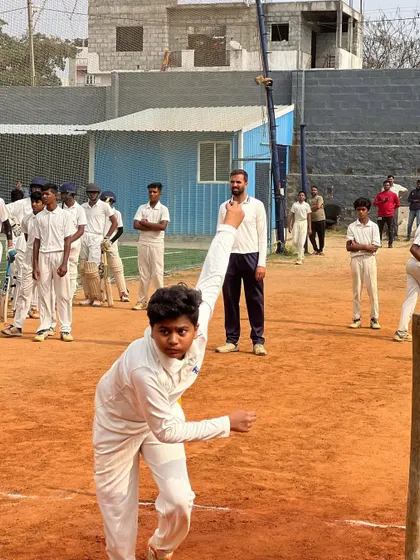 An action shot of a young bowler mid-delivery during an outdoor practice session. The focus and determination are clear as other players watch on.