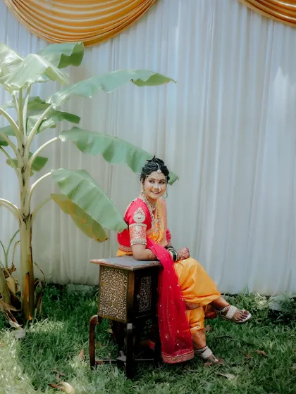 A sweet portrait of a bride in her yellow and pink Nauvari saree, seated outdoors in a natural setting.