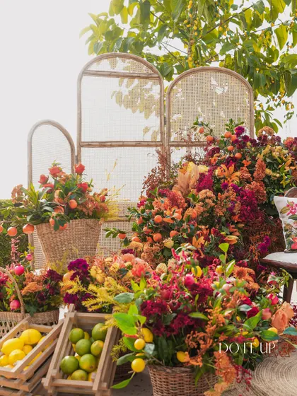 The seating area for the citrus-themed Haldi, featuring a photo-op spot with a cane bench surrounded by an abundance of fresh flowers and fruits like lemons and oranges.