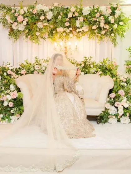 A beautiful Nikkah bride veiled on an elegant white stage, surrounded by pastel pink and white flowers.