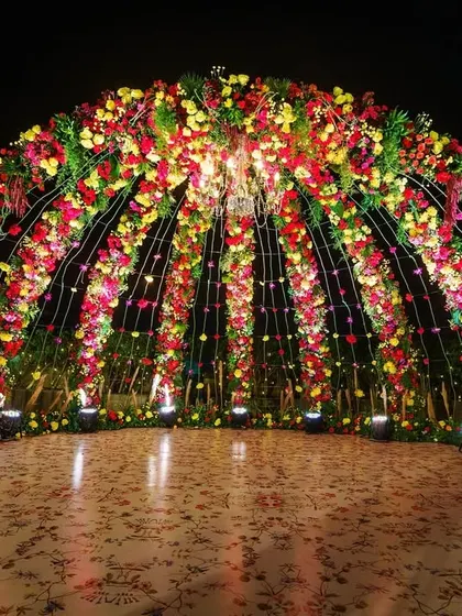 A spectacular floral dome mandap designed for a rooftop wedding. The structure is covered in a vibrant mix of red, yellow, and pink flowers, creating a dramatic and festive atmosphere for the ceremony.