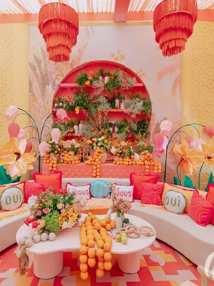 This vibrant seating lounge features a custom-built backdrop with tiered shelves of florals and fresh oranges. We used playful 'Oui' cushions and bold red and orange textiles on clean white sofas to create a fun, high-contrast look.