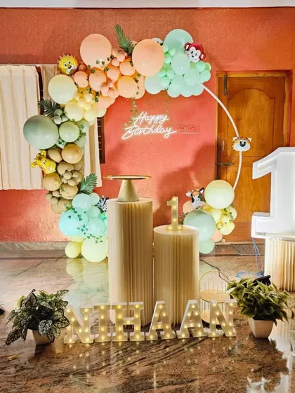 We create modern and gentle themes for first birthdays, like this pastel jungle animal party. The setup includes a soft-colored balloon arch on a ring frame, a 'Happy Birthday' neon sign, large marquee number '1', and illuminated letters spelling the baby's name, 'Nihaan'.