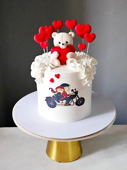 A sweet and simple anniversary cake. This design features a printed edible image of a couple on a motorbike, topped with a cute teddy bear and a halo of red hearts.