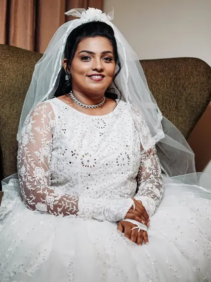 The bride seated, looking calm and beautiful before her ceremony. The full white gown and veil are the stars, supported by a polished and timeless makeup and hair look.