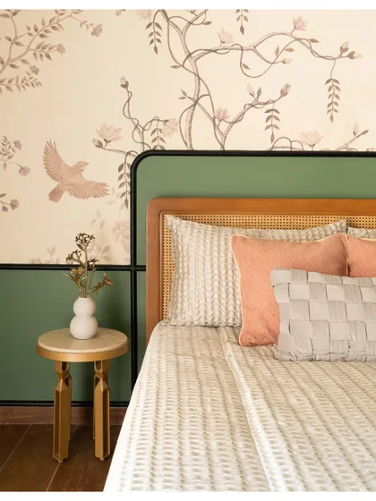 A detail of the bedroom, showing the custom headboard with a green accent panel and a cane inset. The floral wallpaper and gold side table add a touch of elegance.