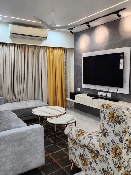A real project photo of a living room in Thane, featuring a grey L-shaped sofa, a floral armchair, and a TV unit against a textured grey wall.