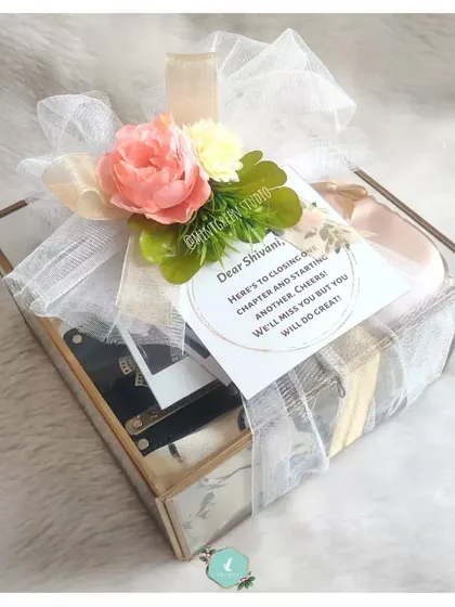 The farewell hamper elegantly packed in a glass and gold-trim box, adorned with a floral arrangement. The personalized note expresses heartfelt wishes for the next chapter, making it a memorable goodbye gift.