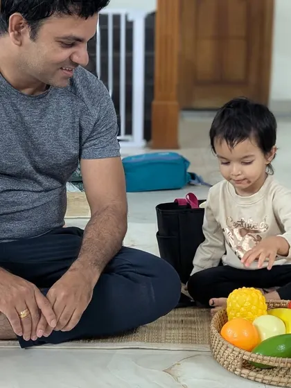 A father watches as his daughter explores a basket of colorful play fruits and vegetables. I encourage parents to be a quiet, reassuring presence as their child begins their journey at Hastam.