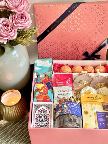 A vibrant pink Diwali box filled with festive delights. This hamper includes Phool incense sticks, playing cards, ladoo candles, choco-coated almonds, and Shubh Labh hangings.