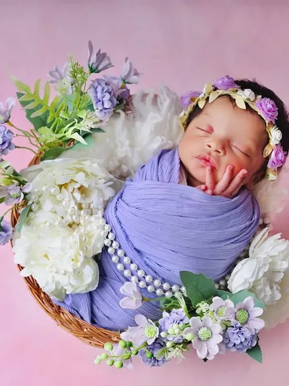 A close-up of a newborn sleeping in a basket, surrounded by a wreath of purple and white flowers. The soft textures and colors create a gentle and artistic portrait.