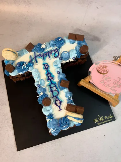 A monogram cake in the shape of the letter "T," decorated in shades of blue with buttercream piping, chocolate pieces, and macarons.
