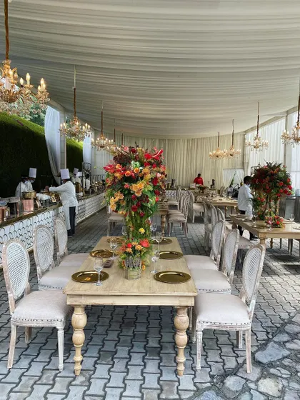 Elegant long-table dining for a sundowner party. The setup features classic wooden tables, upholstered chairs, and stunning, tall floral centerpieces, blending rustic charm with sophisticated design.