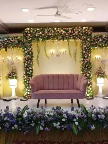 A beautiful engagement stage with a blush pink sofa, framed by a dense arch of pink and white flowers and elegant chandeliers.