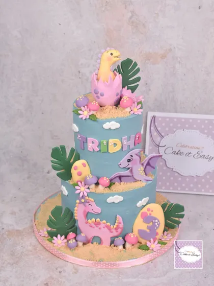 A two-tier dinosaur-themed cake for a little girl's 3rd birthday. The design features cute, friendly dinos in pastel colours, hatching from eggs and surrounded by tropical leaves.