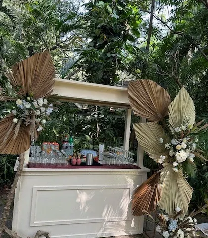 A stylish boho bar cart set up in a lush garden. The cart is decorated with dried palm leaves and white florals, ready to serve refreshing drinks to guests.
