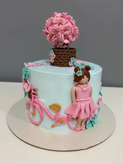 A charming custom cake featuring a 3D fondant girl with her bicycle, surrounded by delicate flowers. The topper is a basket filled with handcrafted sugar flowers.