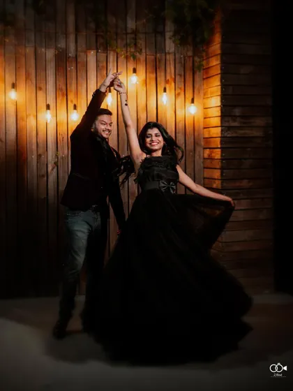 The couple shares a dance under warm string lights, their black outfits creating a classic and romantic evening look.
