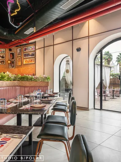 The main dining area at Burosu features long pink leather-upholstered booths and terrazzo tabletops. The arched doorways lead to an outdoor seating area, creating a seamless flow between the indoor and outdoor spaces.