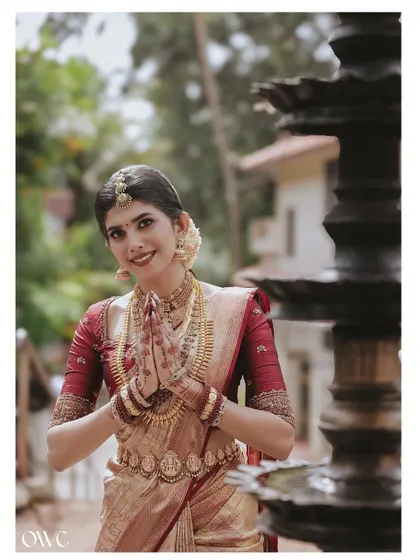 A beautiful portrait of a Kerala bride in a traditional greeting pose, standing by an ornate temple lamp.