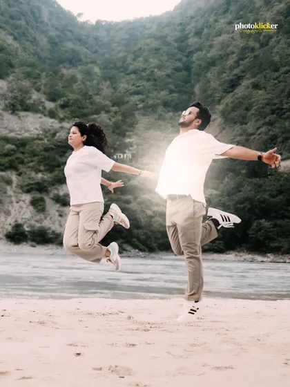 A joyful, high-energy shot of a couple jumping in unison on a river beach, with the mountains providing a stunning backdrop.