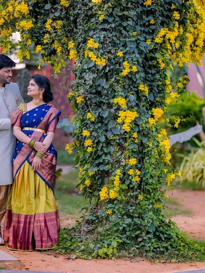 A serene portrait of a couple standing by a tree covered in yellow flowers. Their traditional blue and yellow outfits complement the natural surroundings perfectly.